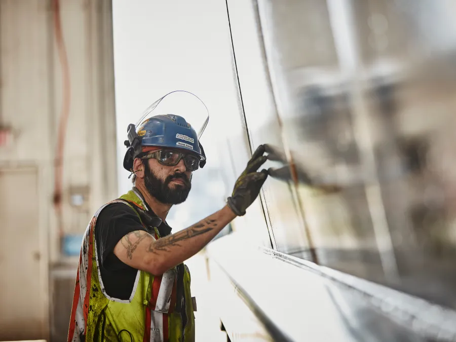A construction worker wearing safety gear and gloves inspects a large reflective metal surface inside an industrial building.