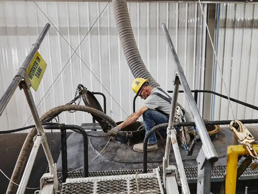 A worker in a hard hat operates industrial machinery, attaching hoses to an open tank in a facility. A "Worker in Tank" warning sign is visible nearby.