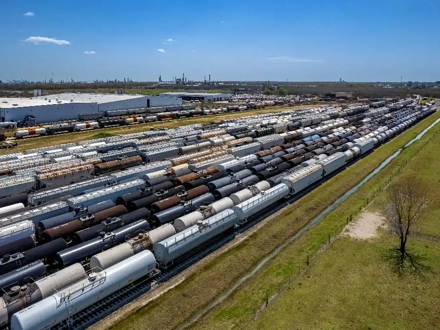 Aerial view of multiple rows of stationary train tanker cars on parallel tracks near a grassy field and industrial buildings under a clear blue sky.