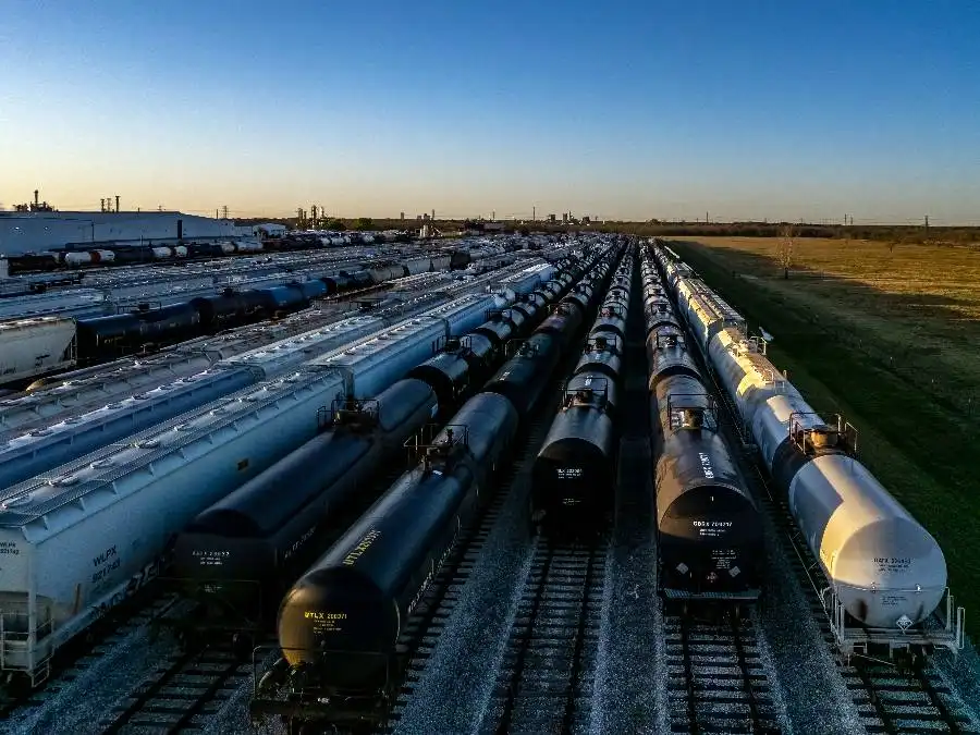 Aerial view of numerous freight train tank cars lined up on parallel tracks in a large rail yard under a clear sky.