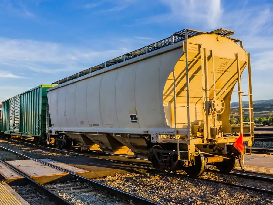 A white covered hopper railcar sits on train tracks next to a green boxcar under a partly cloudy sky.