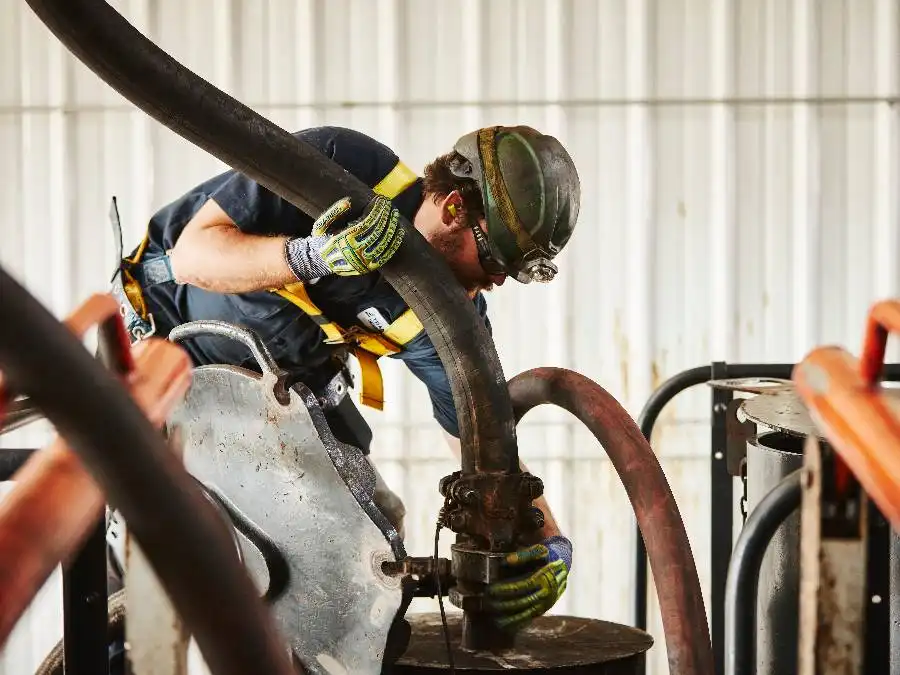 A worker in a hard hat operates industrial machinery, attaching hoses to an open tank in a facility.