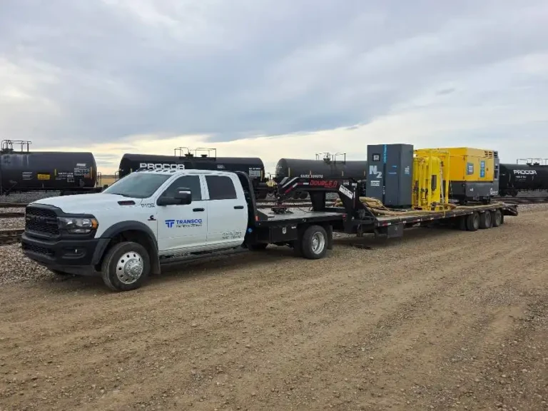 A white pickup truck towing a large flatbed trailer loaded with industrial equipment, parked on a dirt lot near black train tank cars.
