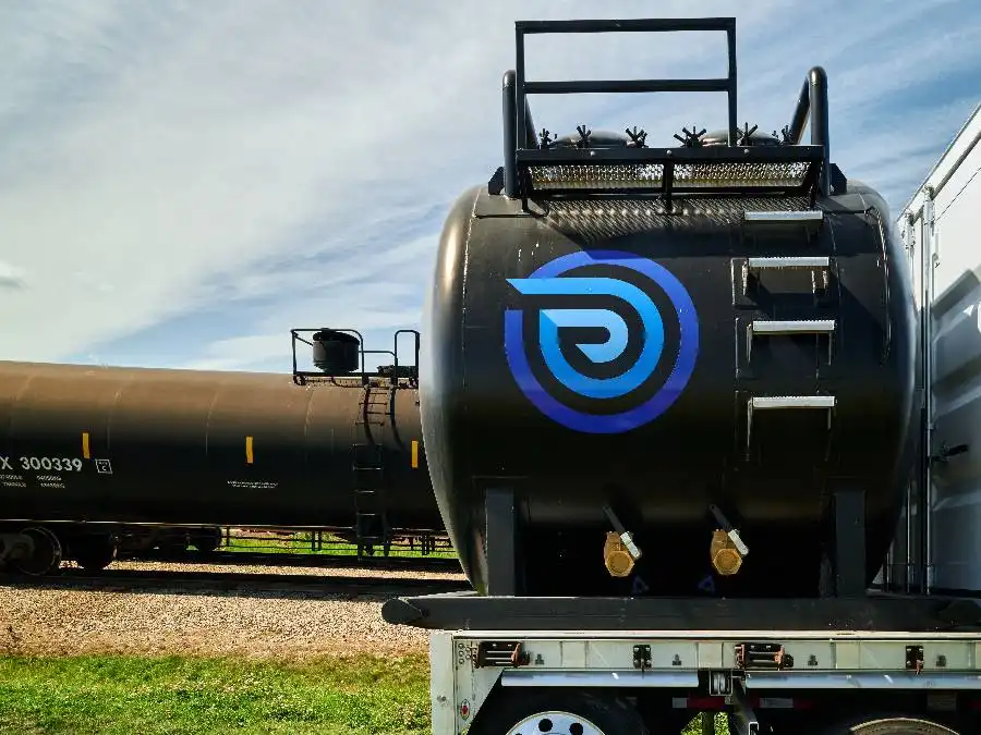 A large black industrial tank with a blue circular logo is mounted on a trailer, with a train tanker car visible in the background.