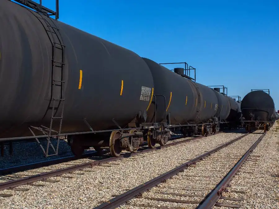 Black cylindrical tanker railcars are parked on parallel train tracks under a clear blue sky.