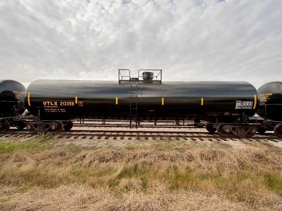 A black cylindrical tanker railcar with the label “UTLX 213359” sits on railroad tracks under a partly cloudy sky, with grass in the foreground.