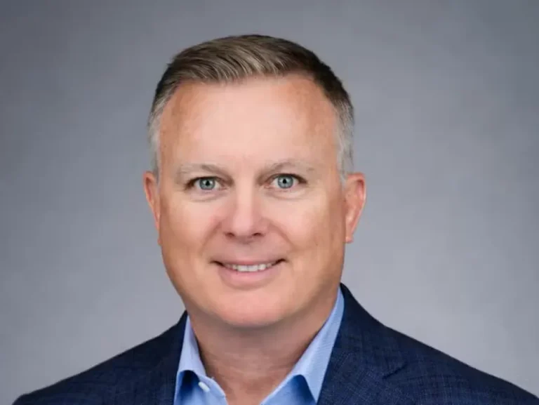 A middle-aged man with short brown hair wearing a blue suit jacket and light blue shirt, posing in front of a plain gray background.