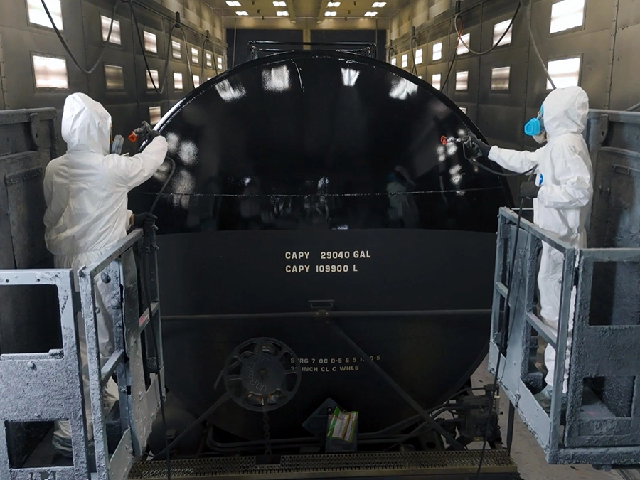 Two workers in protective suits spray paint a large black cylindrical tank inside an industrial facility.