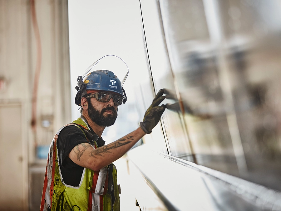 A worker wearing safety gear and a helmet inspects or touches a shiny metal surface inside an industrial facility.