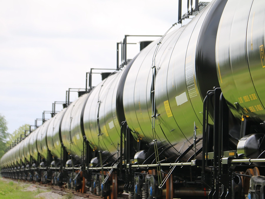 A row of black cylindrical tanker railcars sits on railroad tracks under a cloudy sky.
